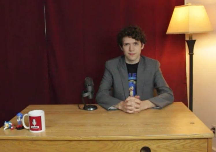 A color photo of Alex Andahazy sitting at a desk with a red curtain behind him. He is white with brown hair, and wearing a gray suit jacket over a Sonic the Hedgehog t-shirt. There is a mug that says 'Talk Hill' on the desk.